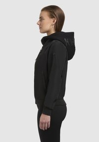 Hoodie zippé noir avec une capuche texturée, arborant un logo au dos, des poignets côtelés et des poches latérales. Le tissu semble lisse et léger.