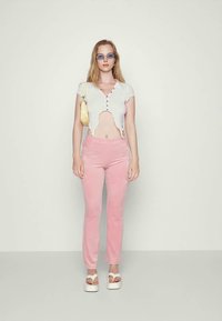 Jovem mulher a usar óculos de sol azuis, uma blusa branca cropped abotoada, calças rosas, sandálias de plataforma brancas e a carregar uma mala de ombro amarela.