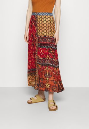 Femme portant une jupe longue patchwork avec des motifs rouges, orange et jaunes, assortie à des sandales beige à boucle et un haut côtelé couleur rouille.