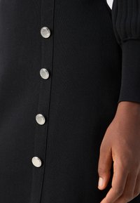 Gros plan sur un vêtement en tissu côtelé noir avec cinq boutons ronds argentés et une main à la peau foncée posée à côté des boutons.