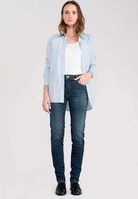 Camisa de botones a rayas azul y blanca sobre una camiseta blanca, combinada con jeans ajustados azul oscuro y zapatos negros, de pie contra un fondo neutro.