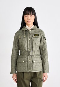 Gequilte olijfgroene jas met een ceintuur in de taille, gouden hardware, vier zakken en een staande kraag. Heeft diamanten patronen en een logo patch.
