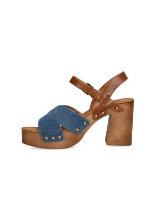 Sandaña marrón con tacón que presenta una tira cruzada de denim. La suela tiene una textura con tachuelas metálicas redondas, añadiendo detalle al diseño general.