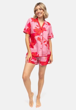 Kobieta stojąca boso, ubrana w różową koszulę na guziki z krótkim rękawem i krótkie spodenki z dużym czerwonym kwiatowym wzorem, uśmiechająca się i patrząca lekko w lewo.