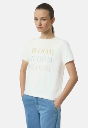 T-shirt en coton blanc avec "BLOOM" imprimé en lettres colorées et en pointillés. Col arrondi et manches courtes. Le modèle porte un jean bleu clair.