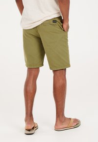 Olivgrüne Shorts aus Baumwolle. Mit einer Gesäßtasche mit Branding. Kombiniert mit beigen Flip-Flops, präsentieren sie die Beine und bieten eine lässige Passform.