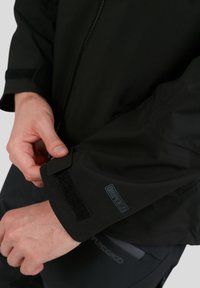 Zwarte waterdichte jas met een klittenbandsluiting bij de mouwen en een rubberen logo patch op de mouw, met een gladde, matte textuur.