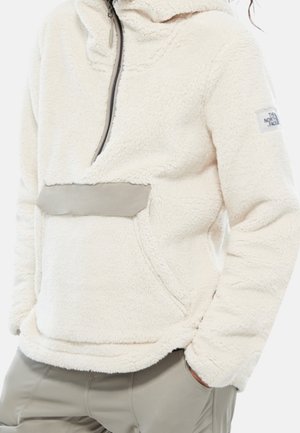 Osoba w białym polarku z półzamkiem, z kieszenią z przodu i logo The North Face na lewym rękawie.