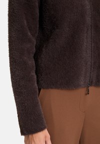 Giacca zip-up a cropped design di un marrone scuro e morbido. Texture soffice, maniche lunghe e dettaglio della zip in metallo. Abbinata a pantaloni marroni.