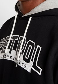 Sudadera negra con interior gris, que presenta un prominente logo blanco. El diseño incluye cordones y letras texturizadas.
