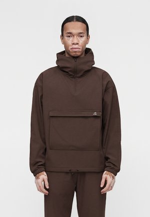 Man with vitiligo wearing a brown high-neck pullover with large front pocket and matching pants against a plain light background.