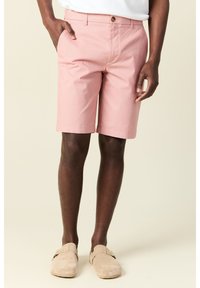 Vaaleanpunaiset puuvillashortsit litteällä etuosalla, yhdessä ruskealla napilla kiinnitys, sivutaskut ja rento malli, yhdistettynä vaaleisiin liukuvaan kenkään.