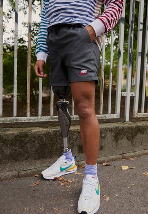 Person trägt schwarze Nike-Shorts, ein robotisches Prothesen- linkes Bein, weiße Nike-Sneakers und ein gestreiftes Langarmshirt nahe einem Metallzaun.