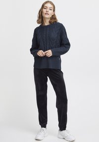 Marinblå stickad tröja med kabelmönster, ribbade ärmslut och nederkant; matchad med svarta sammetsbyxor och vita sneakers. Enkel och avslappnad outfit.
