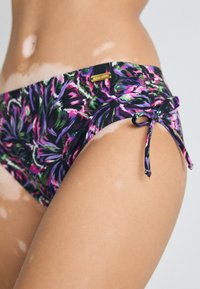 Floral-patterned bikini bottom featuring shades of purple, green, and pink, with adjustable side ties and a small brand tag.