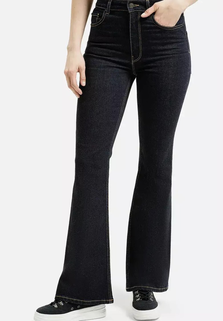 Kobieta w czarnych, wysokich dżinsach w stylu flare i czarnych sneakersach, stojąca z jedną ręką w kieszeni, a drugą swobodnie przy boku.