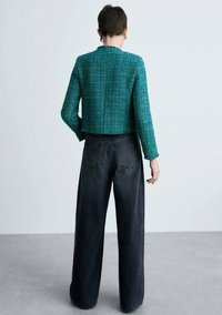 Giacca in tweed verde acqua tagliata, con superficie testurizzata, abbinata a jeans neri a gamba larga con tasche posteriori e una leggera sfumatura.