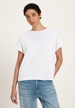 Frau mit kurzen, lockigen Haaren, die ein schlichtes weißes T-Shirt mit kurzen Ärmeln und hellblaue Jeans trägt, vor neutralem Hintergrund stehend.