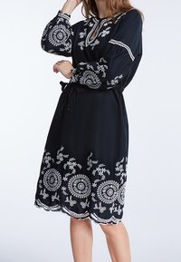 Robe noire à manches longues, broderie florale blanche, taille cintrée et ourlet en points de feston. Présente un décolleté en V et une texture de tissu douce.