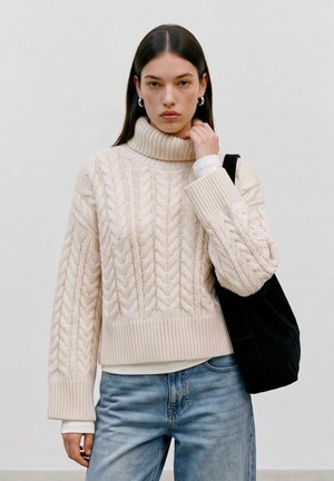 Femme aux longs cheveux foncés portant un pull à col roulé en maille torsadée crème, un jean bleu clair, des boucles d'oreilles anneaux argentées et tenant un sac à bandoulière noir.