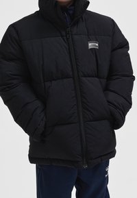 Personne portant une doudoune noire avec les mains dans les poches et un pantalon bleu marine sur un fond clair uni.