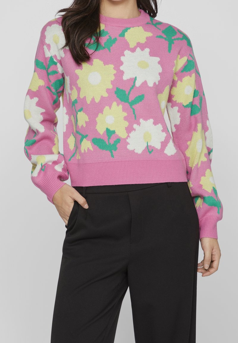 Suéter rosa de punto con grandes patrones florales blancos y amarillos, con hojas verdes, y puños y dobladillo acanalados. Combinado con pantalones negros.