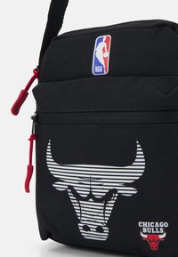 Melnā sporta soma ar strukturētu virsmu, ar baltu svītrotu Čikāgas Bulls logo un mazu NBA logo, sarkano rāvējslēdzēju velcējs.