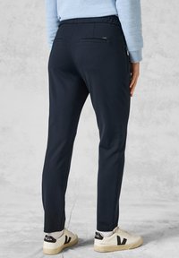 Mörkblå slim-fit joggers med elastisk midja, två bakfickor och en mjuk textur, tillsammans med vita och svarta sneakers.