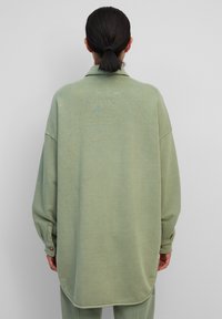 Persoon met donker haar in een lage paardenstaart draagt een oversized lichtgroen knoopshirt en bijpassende broek, staand tegen een effen achtergrond.