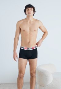Caleçons boxers pour hommes noirs avec un logo "DIESEL" blanc et rouge sur la ceinture. Le modèle se tient dans un cadre neutre, les mains sur les côtés.