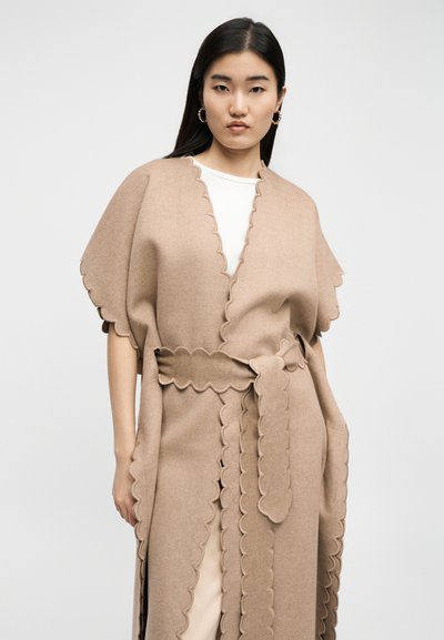 Γυναίκα με μακριά μαύρα μαλλιά φοράει μπεζ παλτό με κυματιστές άκρες πάνω από λευκό τοπ, ποζάροντας μπροστά σε απλό ανοιχτόχρωμο φόντο.