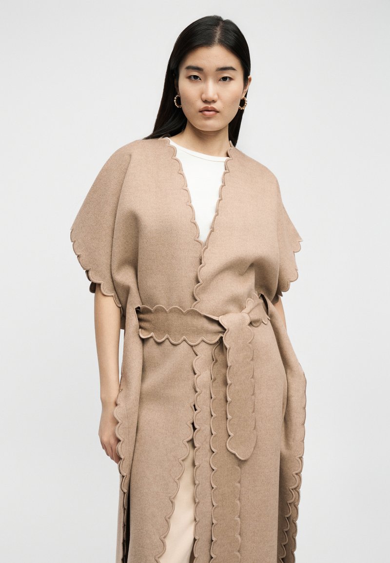 Frau mit langen schwarzen Haaren trägt einen beigen Wickelmantel mit wellenförmigem Saum über einem weißen Oberteil und posiert vor einem schlichten hellen Hintergrund.