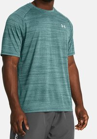 Teal lyhythihainen urheilupaita, jossa on tekstuurikuvio ja pyöreä kaula-aukko. Vasemmassa rinnassa pieni valkoinen logo.