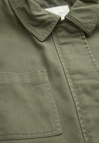 Camisa verde oliva con cuello doblado, un bolsillo en el pecho izquierdo y tejido texturizado con detalles de costura visibles.