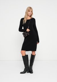 Schwarzes Strickkleid mit langen Ärmeln, geripptem Saum, kombiniert mit knielangen schwarzen Lederstiefeln und einer kleinen schwarzen Handtasche.
