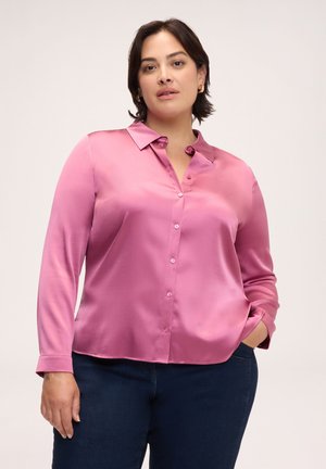 Kvinde iført en skinnende lyserød skjorte med knapper og lange ærmer samt mørkeblå jeans, stående med den ene hånd i lommen mod en ensfarvet baggrund.