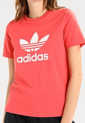 Person iført en koralfarvet Adidas t-shirt med hvid trefoil-logo og tekst, kombineret med sorte bukser, vist fra nakke til hofter.