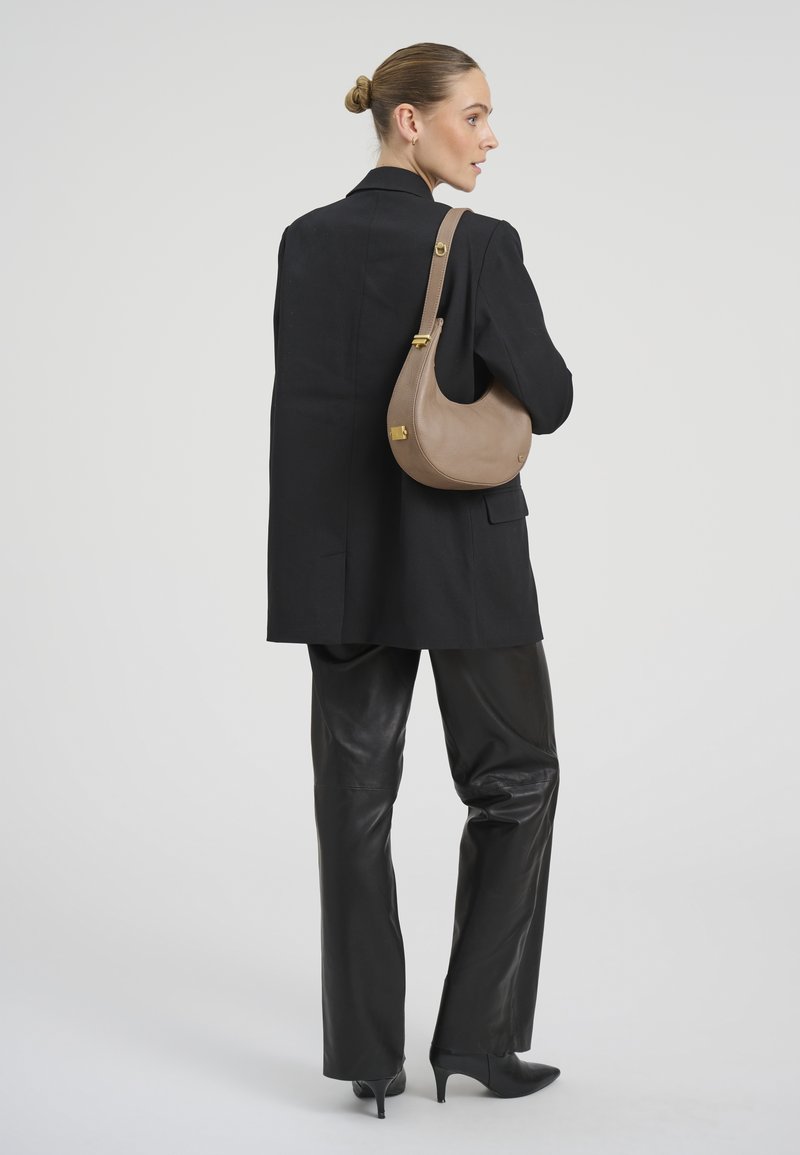 Femme avec chignon porte un blazer noir, un pantalon en cuir, des talons et un sac à bandoulière beige, debout de profil sur fond gris.