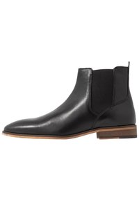 Černá kožená Chelsea bota s elastickými bočními panely, špičatou špičkou a světle hnědým vrstveným podpatkem. Hladká textura s minimálními detaily.