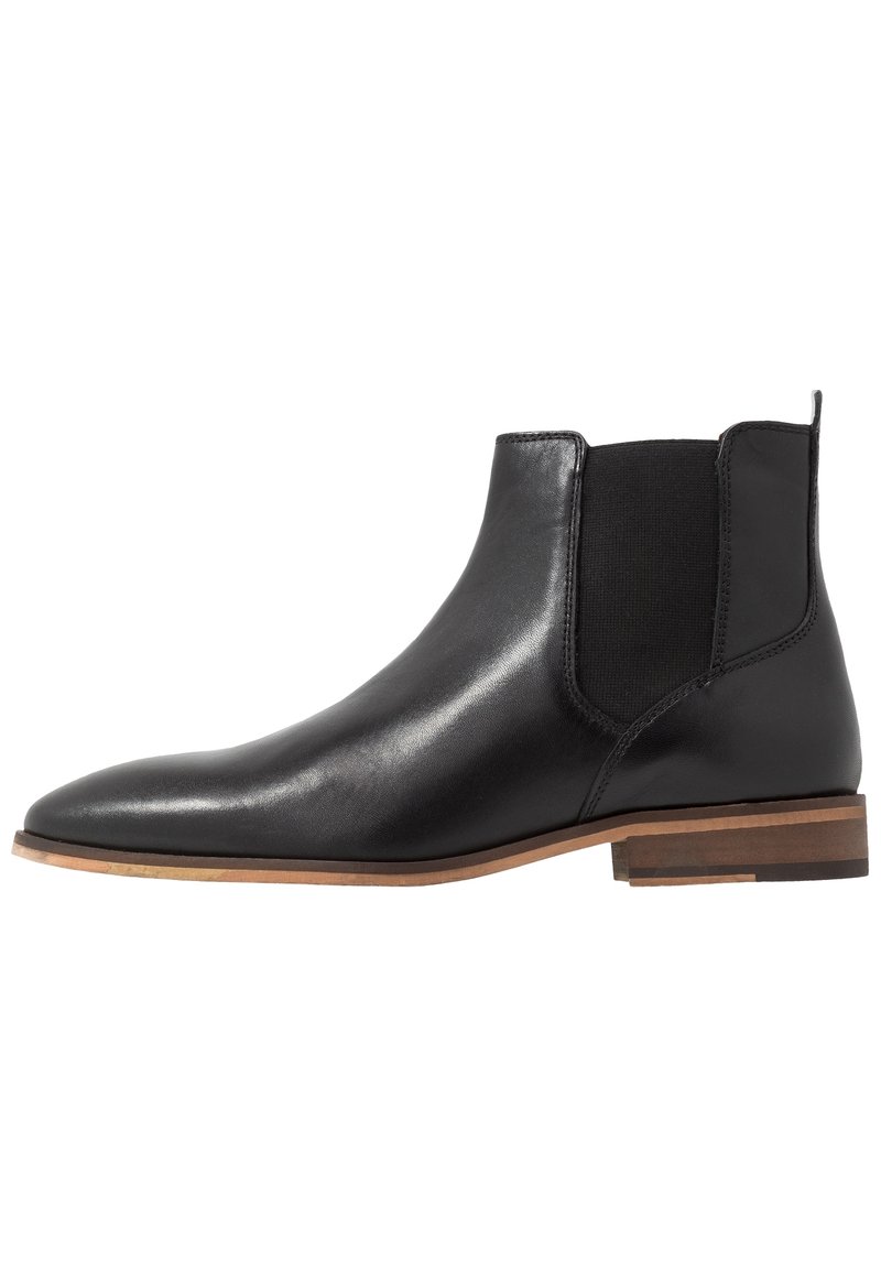 Černá kožená Chelsea bota s elastickými bočními panely, špičatou špičkou a světle hnědým vrstveným podpatkem. Hladká textura s minimálními detaily.