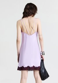 Una donna con i capelli castani mossi indossa un vestito slip lavanda con spalline sottili e bordo in pizzo scuro, mentre tiene una borsa nera e bracciali d'argento.