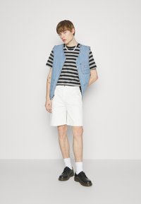 Denimvest med åben front, sort og hvid stribet t-shirt, hvide shorts og sorte sko. Har et enkelt design og en afslappet pasform.