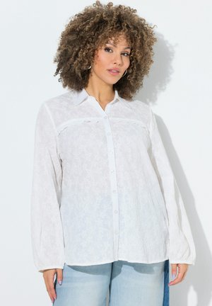 Frau mit lockigem Haar, die eine weiße, mit Blumen bedruckte Bluse mit Knöpfen und hellblaue Jeans trägt, steht vor einer weißen Wand.