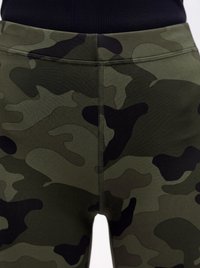 Close-up van groene en zwarte camouflage patroon leggings gedragen met een zwarte top, met details van stiksels en tailleband.
