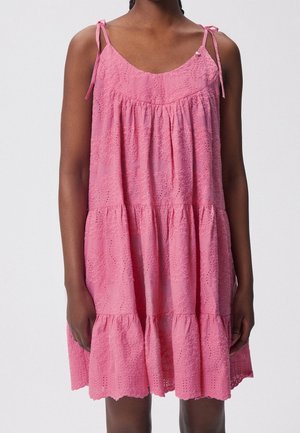 Vrouw draagt een mouwloze roze jurk met meerdere lagen, oogjesborduursel en gestrikte schouderbandjes, staand tegen een effen achtergrond.