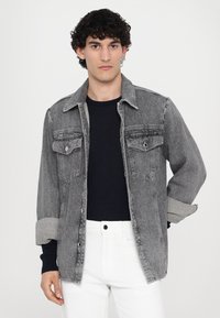 Gray denim jacket with two chest pockets, rolled sleeves, and a straight cut, layered over a navy long-sleeve shirt and white pants.