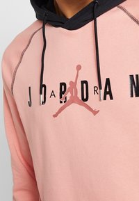 Hoodie rosa chiaro con dettagli neri e un cordino. Presenta un grande logo nero "JORDAN" e un grafico in silhouette di un rosa più scuro.