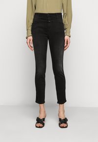 Vaqueros de mezclilla negra con un corte ajustado, cintura de altura media y un desvanecimiento sutil. Combinados con una blusa verde holgada y sandalias negras con nudo.