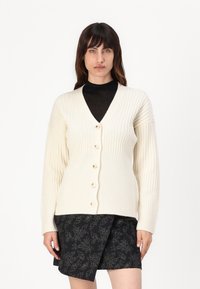 Cardigan a coste color crema con maniche lunghe, caratterizzato da un collo a V, sei bottoni beige sul davanti, abbinato a una gonna nera a fantasia.