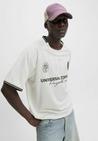 Camisa blanca de manga corta con detalles en negro, que presenta el texto "ESTÁNDARES UNIVERSALES" y un logotipo circular. Combinada con pantalones cortos de mezclilla azul claro y una gorra morada.
